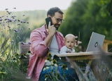 Man working on a laptop and talking on a phone while holding a baby
