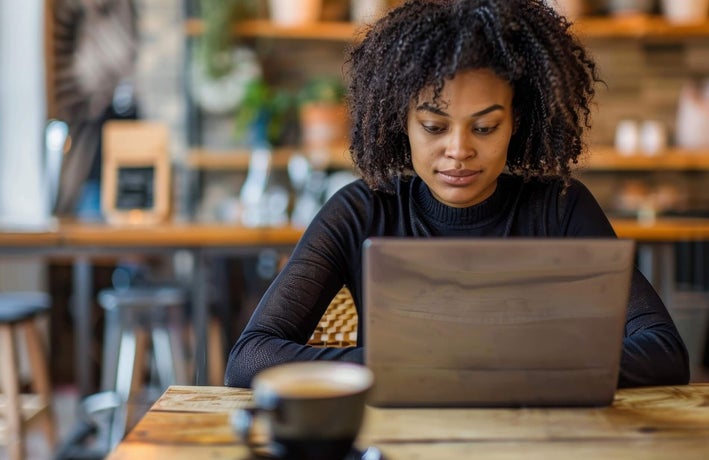 A woman with a black sweater sitting at wooden cafe table with a brown laptop.