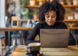 A woman with a black sweater sitting at wooden cafe table with a brown laptop.